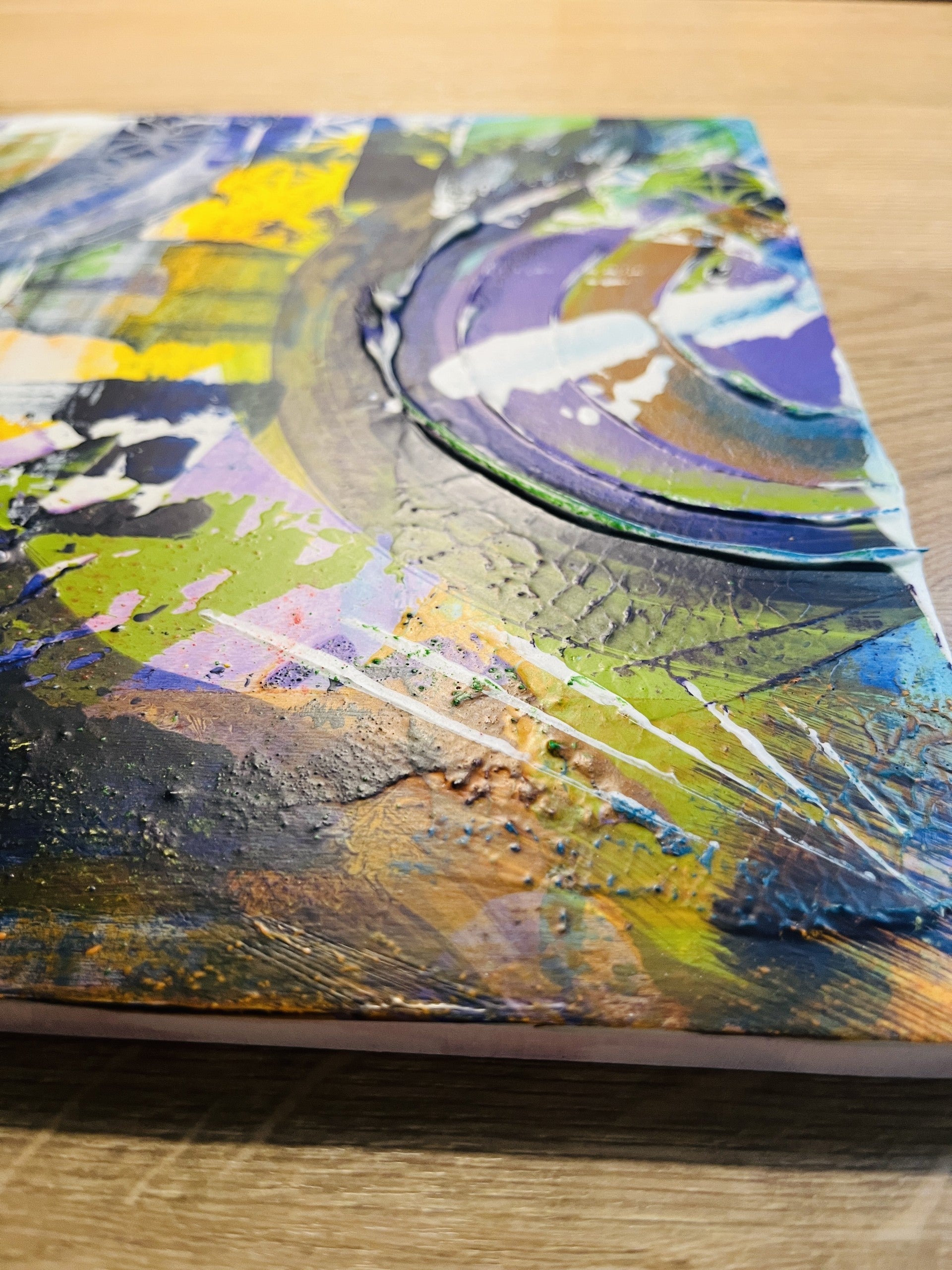 An abstract textured painting on canvas, featuring thick impasto brushstrokes in purple, green, yellow, blue, black, and white, resting on a light wood surface.