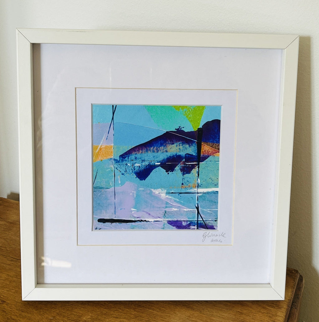 A framed abstract art print featuring a colorful fish illustration, displayed on a wooden table against a white wall.