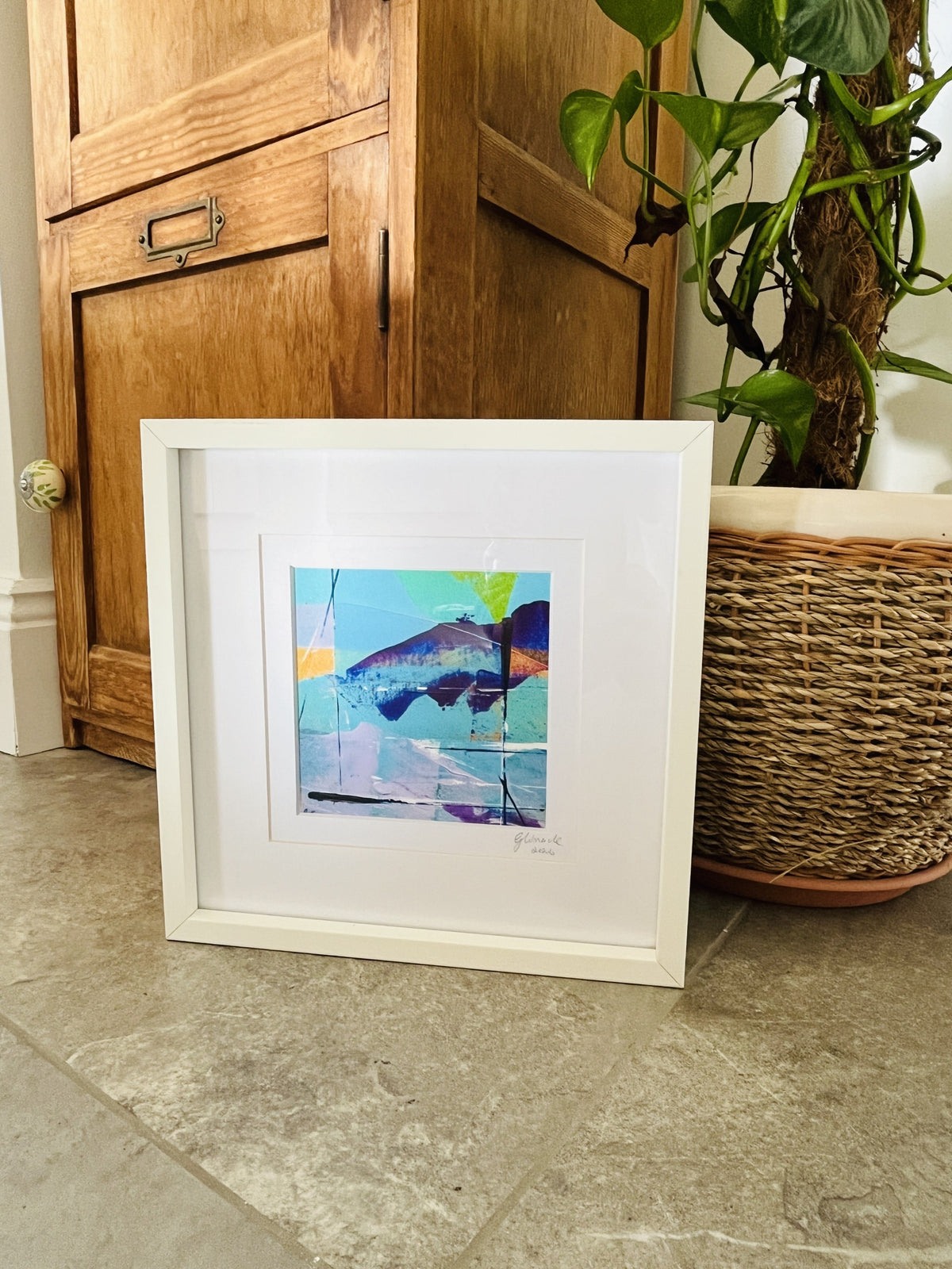 A framed abstract art print featuring a mountain landscape in shades of blue, purple, and green, set in a white frame on a stone floor next to a wooden cabinet and a potted plant.