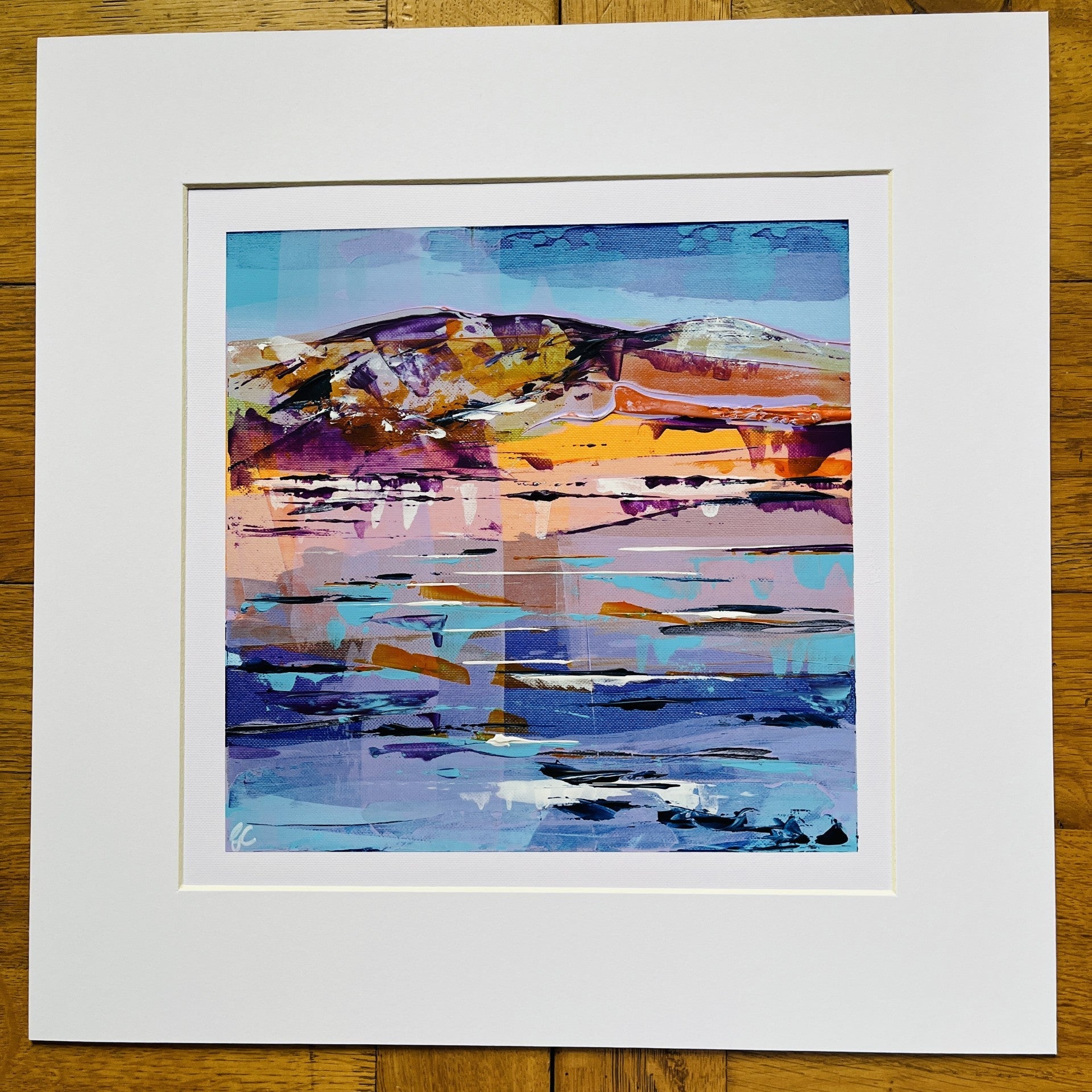 An abstract landscape art print with vibrant blue, purple, orange, and yellow brushstrokes, presented with a wide white mat board on a rustic wooden background.