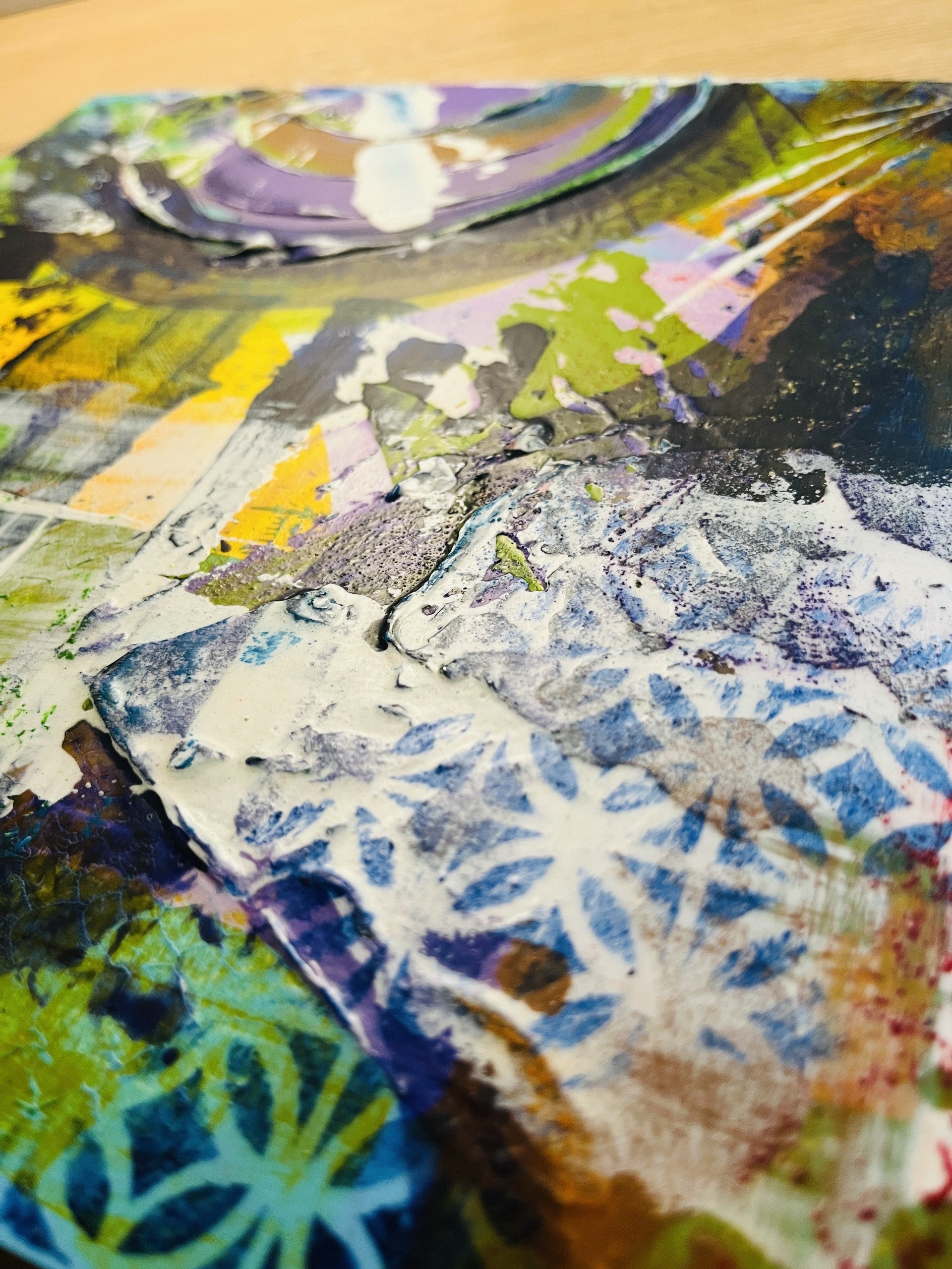 A close-up of a mixed-media art print or textured collage featuring abstract patterns in blue, purple, green, yellow, black, and white, with a glossy finish.