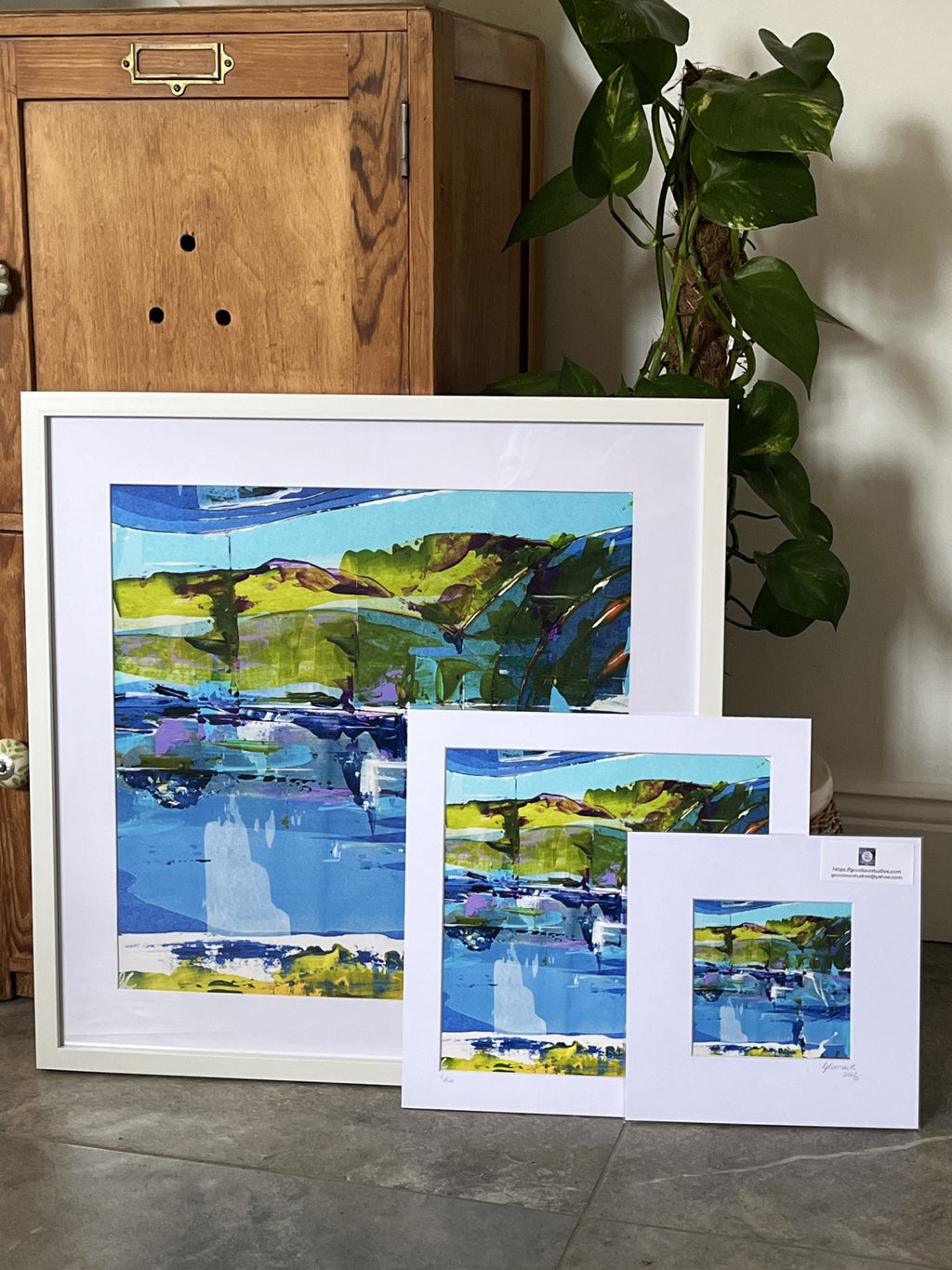 Three framed landscape art prints in different sizes, featuring an abstract painting of blue water, green hills, and purple mountains, displayed on a gray floor next to a wooden cabinet and a green plant.