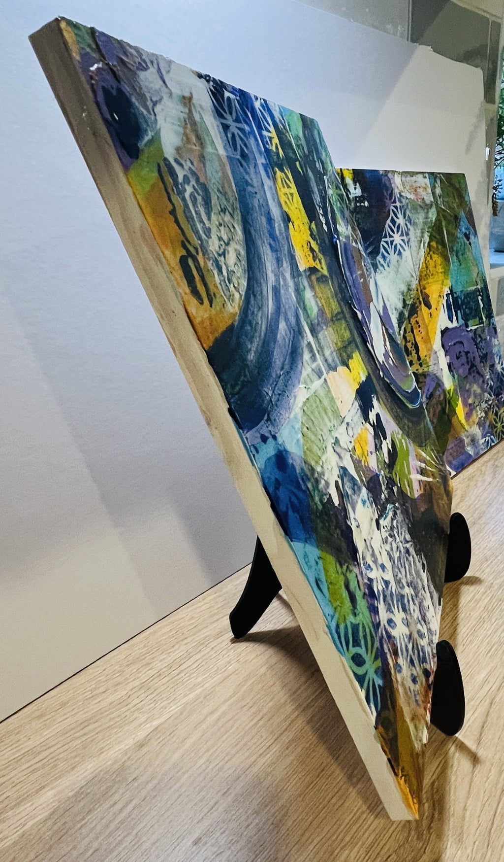 Abstract mixed-media painting on a square wooden board, displayed standing on a black easel in an indoor setting with a light-colored wall and wood floor.