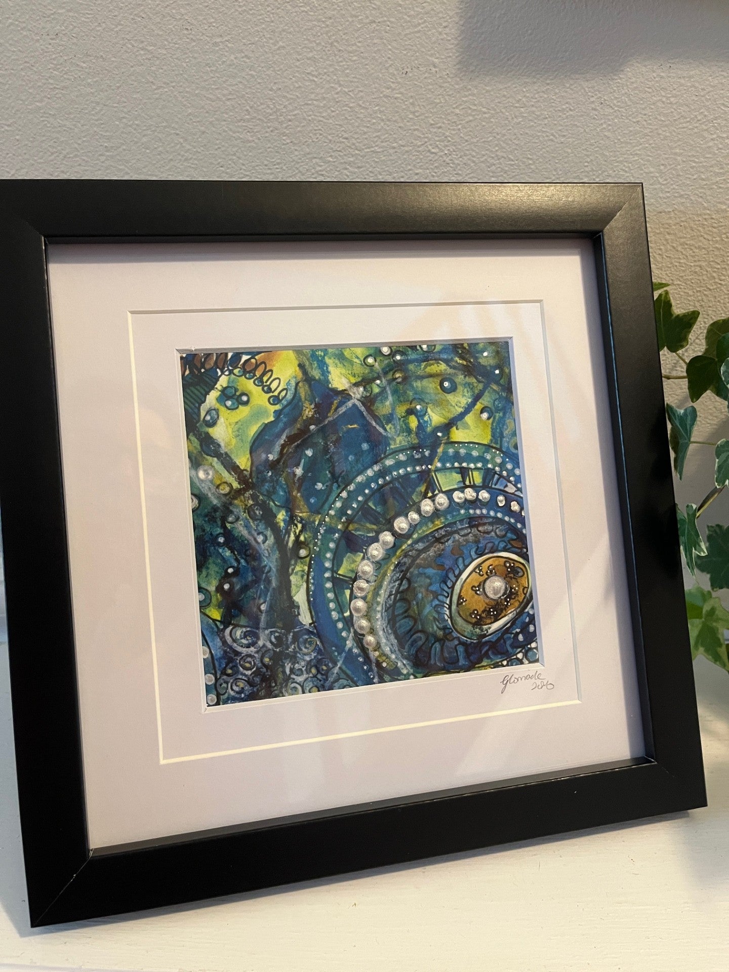 Framed abstract artwork with blue and green colors on a white surface.