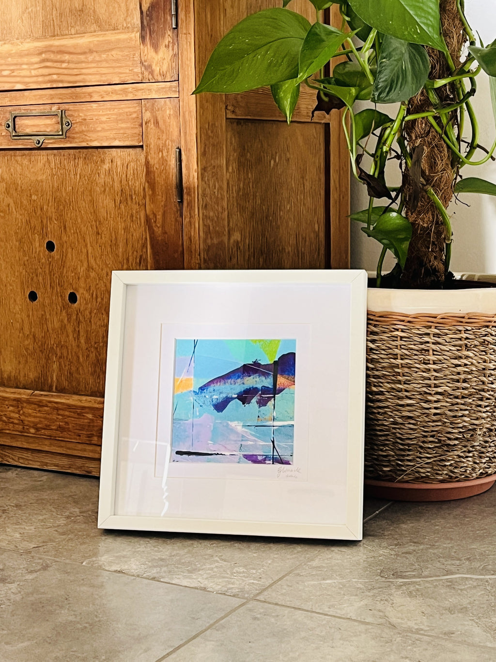 A framed abstract art print with a white frame and mat, leaning against a wooden cabinet on a tiled floor next to a potted green plant.